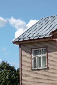A residential metal roof with snow partially melted off, set against a blue sky and a few nearby trees.