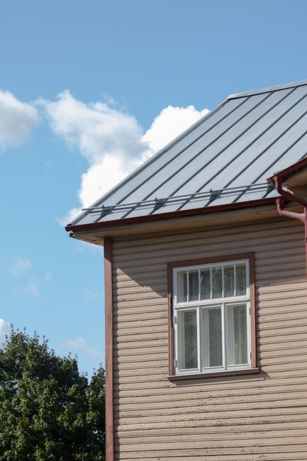 A residential metal roof with snow partially melted off, set against a blue sky and a few nearby trees.