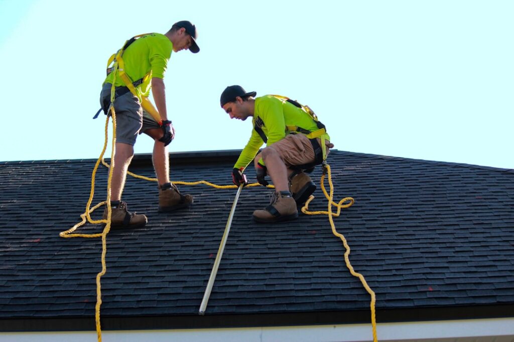 A person in safety gear examines asphalt shingles on a sloped residential roof under a bright sky.